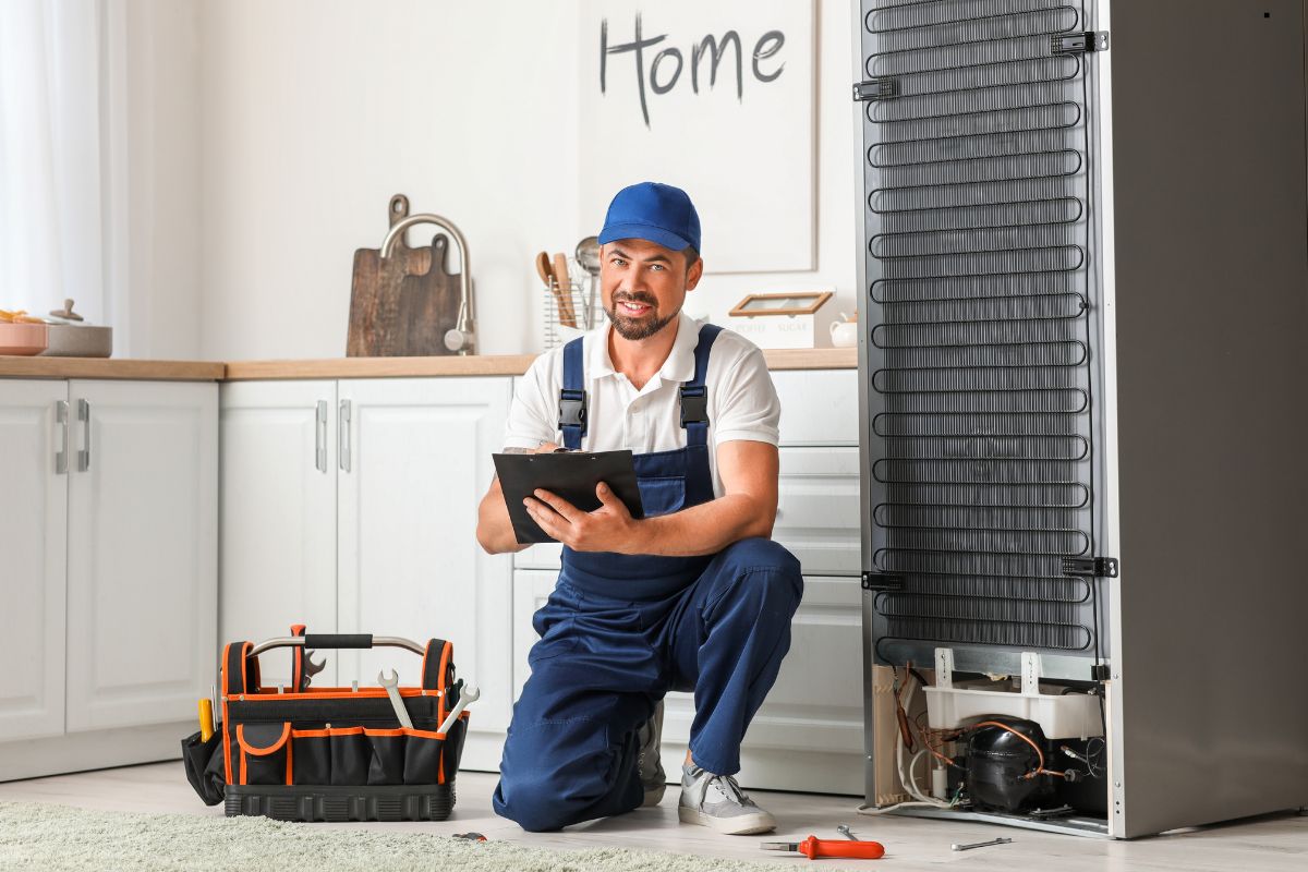 A man wearing overalls is seated on the floor next to a toolbox and a refrigerator, focused on his work.