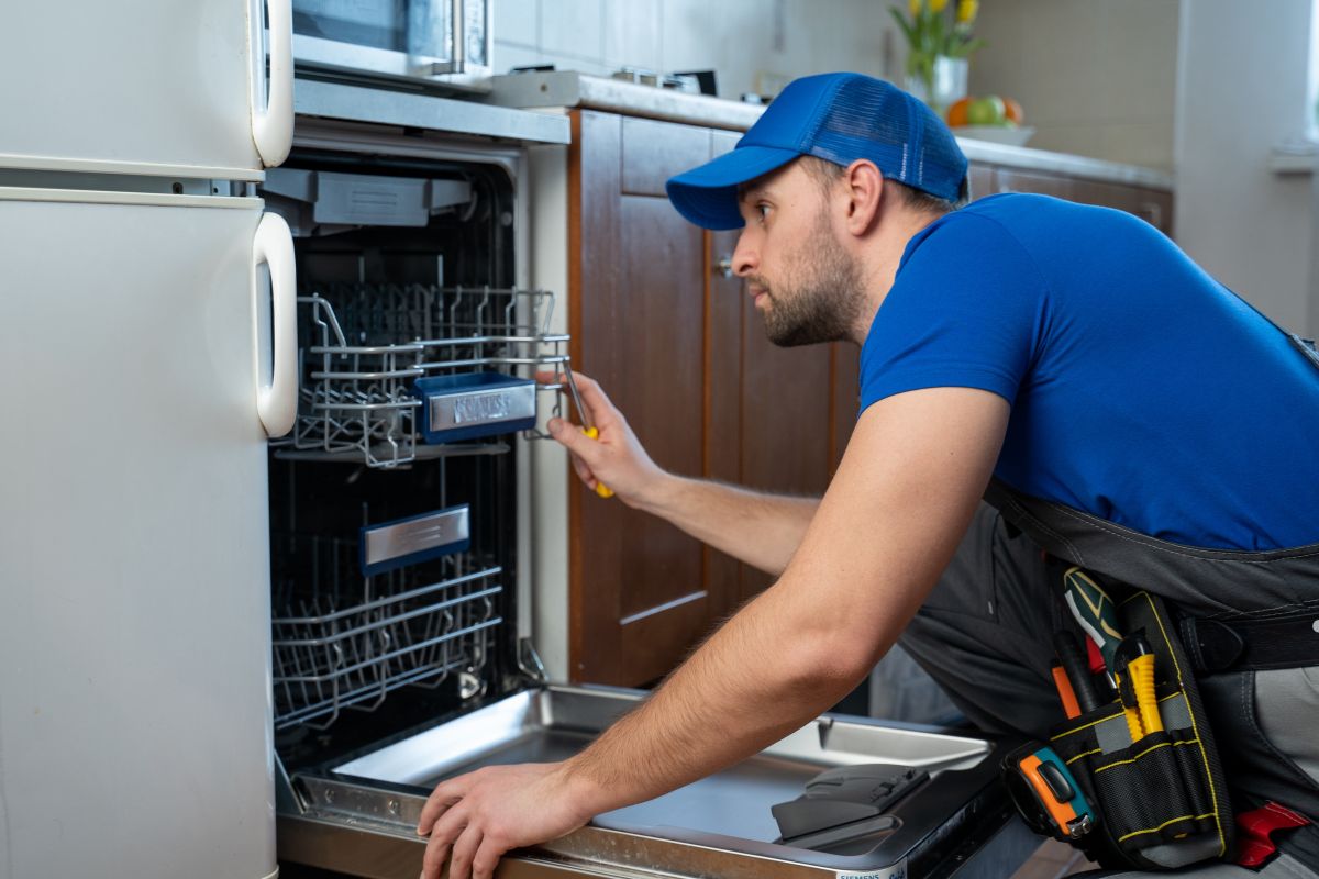 A technician in a blue shirt and hat repairs a dishwasher, showcasing appliance repair service expertise.